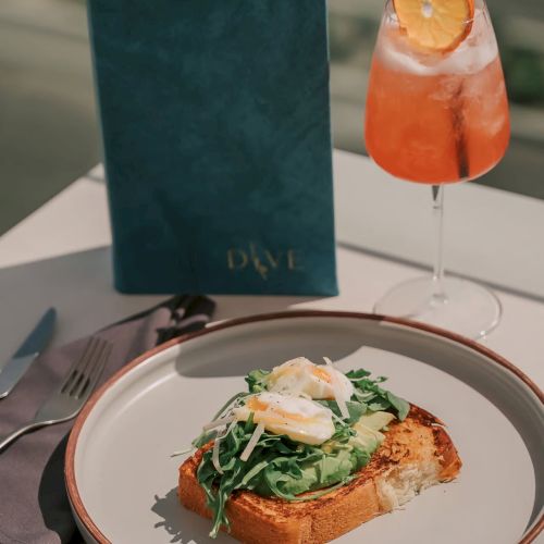 A plate with toast topped with greens and poached eggs, next to a drink with an orange slice and a blue menu at the back.