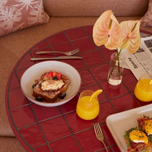 A cozy table setup with brunch items: plates of food, orange juice, anthurium flowers in a vase, and a sofa with floral cushions.