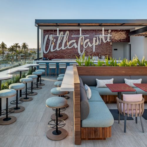 Rooftop lounge with modern seating, tables, and a large "Villa Capri" sign on a brick wall. Palm trees visible in the background.