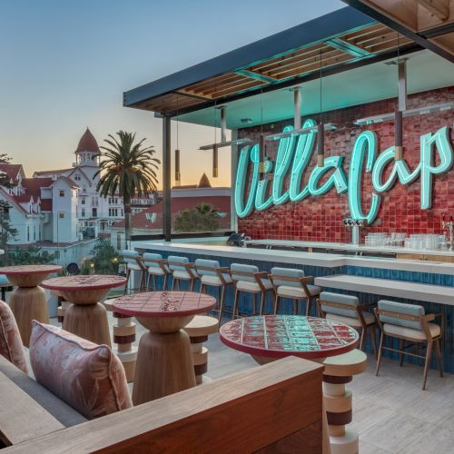 The image shows a stylish outdoor bar area with "Villa Capri" in neon, set against a backdrop of palm trees and a large building.