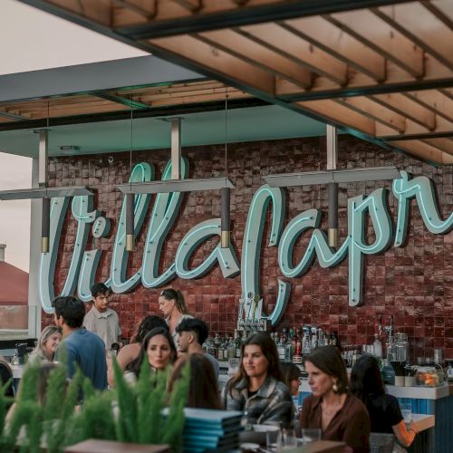 People are gathered at a bar or restaurant with a neon sign reading "Villa Capri" in the background.