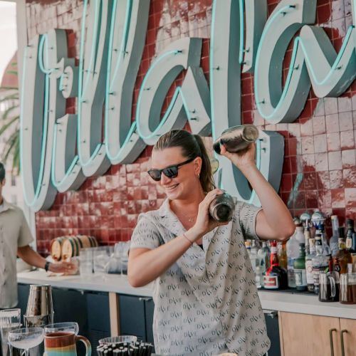 A bartender in sunglasses is shaking a cocktail shaker at a bar with a "Villa" sign in the background.