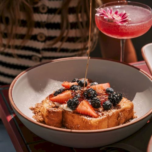 A plate of French toast with strawberries and blackberries, drizzled with syrup, beside a pink cocktail with a flower garnish.