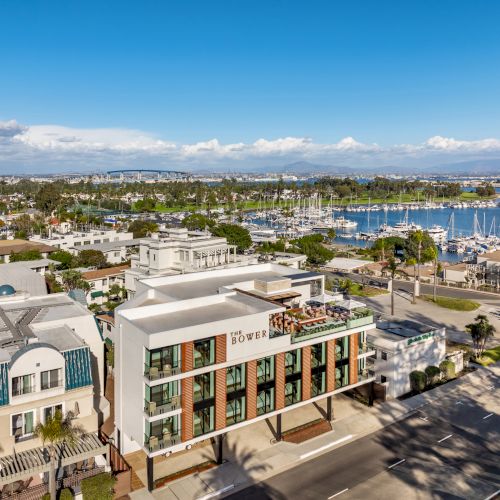 Aerial view of a coastal area with a modern building, marina, and sailboats, set against a backdrop of blue sky and distant hills.