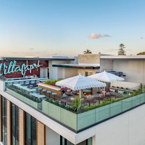 A rooftop terrace with seating, umbrellas, and a sign that reads "Villa Capri" on a modern building under a clear blue sky.