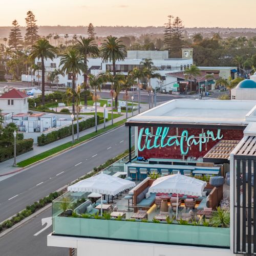 A rooftop terrace with umbrellas and seating overlooks a street with palm trees, near the coast, at sunset, with buildings and greenery nearby.