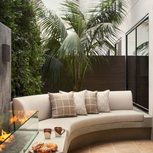Modern outdoor patio with a curved sofa, pillows, a fire feature, palm plants, and snacks on a small table, creating a cozy ambiance.