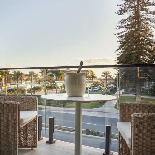 A small round white table on a balcony with a metal railing, overlook of trees and a city/valley beyond, sunny day.