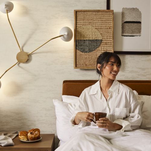 A man sits up in bed, reading a book, with a wall clock and framed art behind him, a bedside table with a plant or lamp nearby.