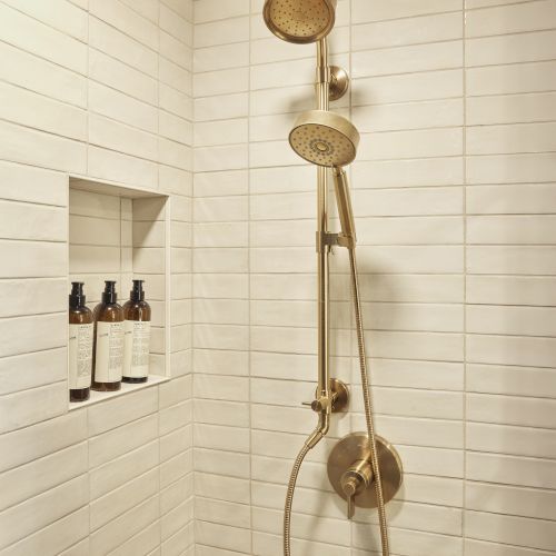 A modern shower with a tall rainhead and a handheld wand, plus a small set of wall-mounted shelves holding bottles.