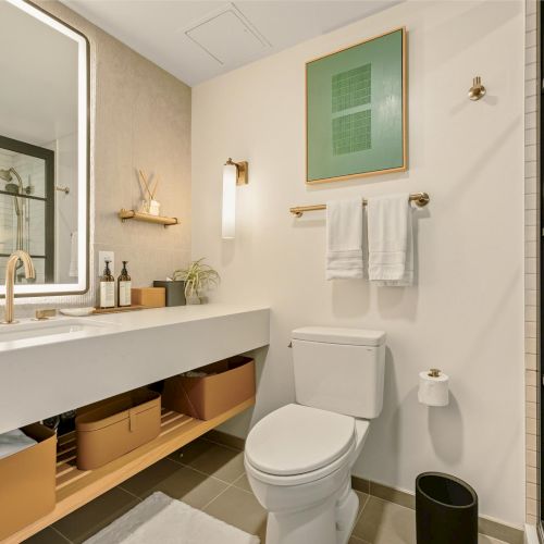 A modern bathroom with a white double-sink vanity, large backlit mirror, open storage shelves, wall-mounted towels, toilet, and a glass shower enclosure.