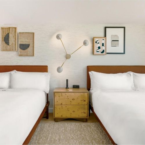 Two neatly made hotel rooms with two twin beds each, a shared nightstand between them, and minimalist wall art above a textured lamp on the left side.