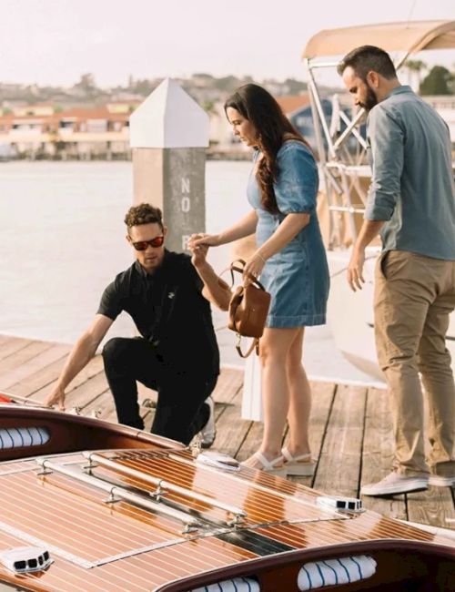 A couple and a man are at a dock with a wooden speedboat, engaging in conversation, while the boat is moored.