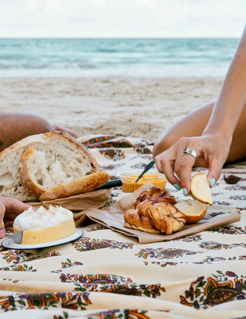 A beach picnic scene with two people, bread, pastries, fruit, and desserts on a patterned blanket by the shore.