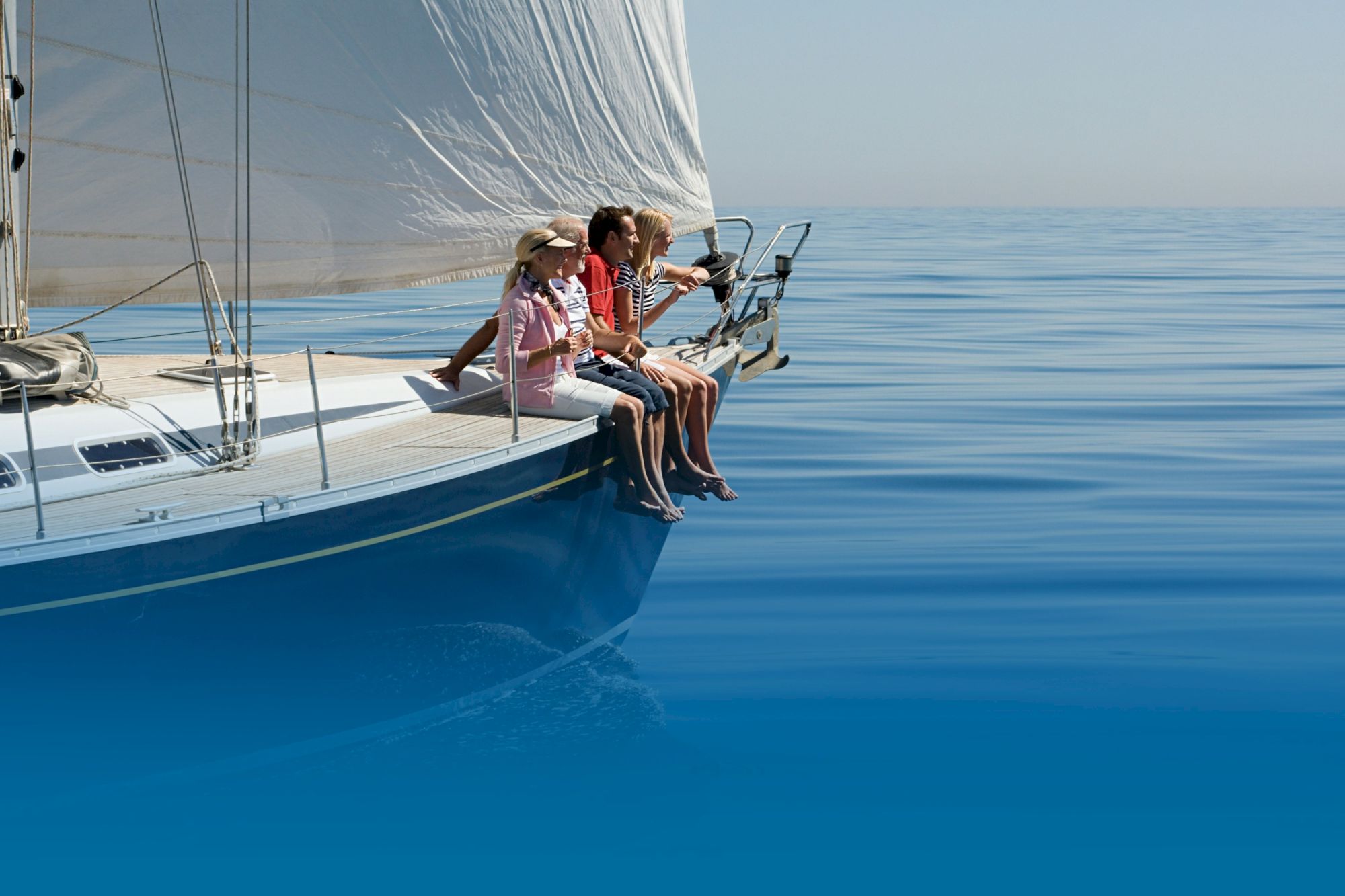 A group of people sitting on the edge of a sailboat, enjoying a calm and clear day on the serene blue sea.