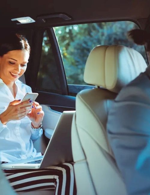 A woman in the backseat uses her phone, while a man drives the car. The interior looks modern and comfortable.