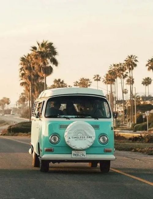 A vintage teal van drives on a sunny road lined with palm trees.