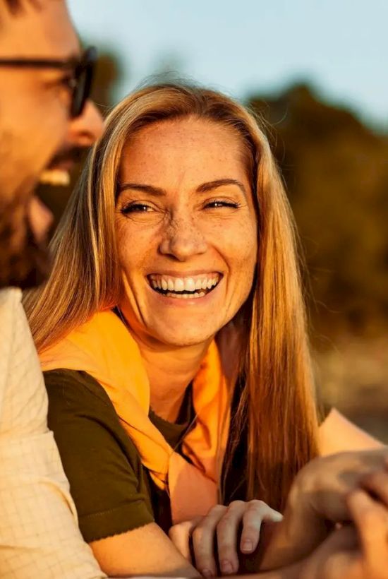 A man and woman are outdoors laughing, with trees and a warm sunset in the background.