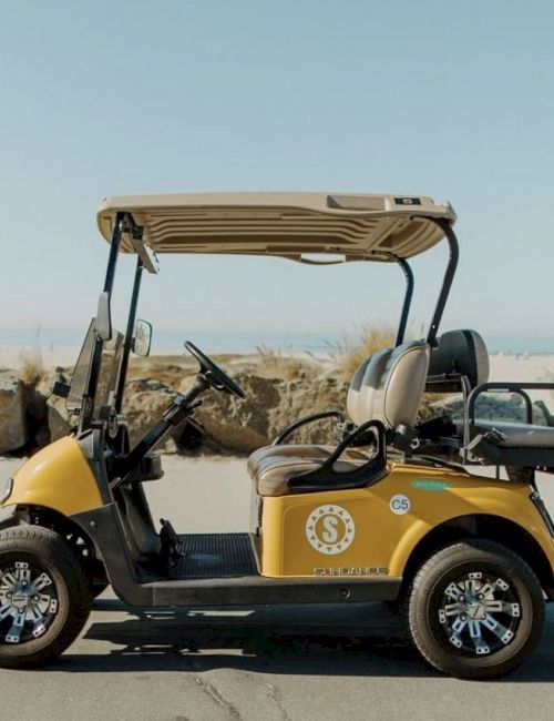 A yellow golf cart with a beige canopy parked on a sidewalk near rocky coast, logo on the door, black seats, and chrome wheels, sunny day.