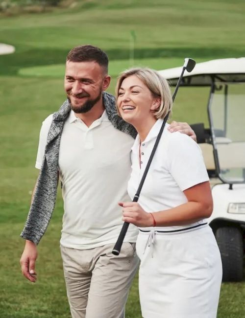 A smiling man and woman stand on a golf course with a cart in the background, she holds a club, both dressed casually in sports attire.
