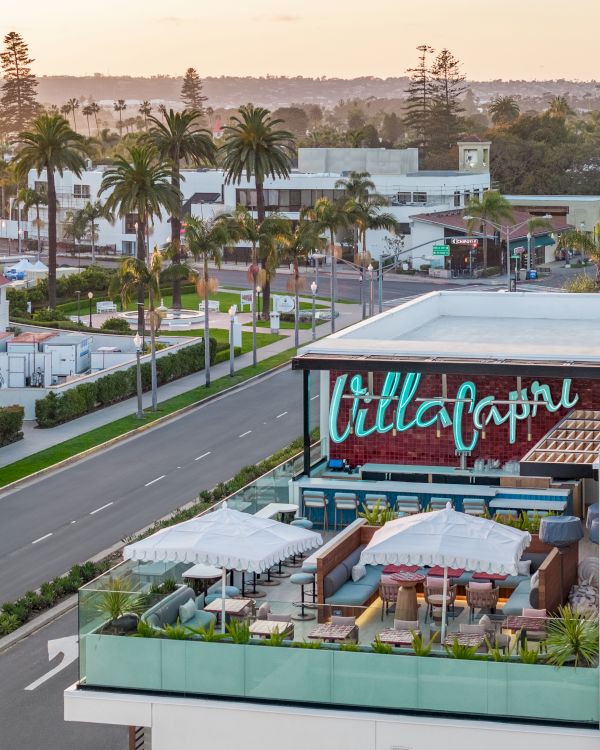 A rooftop lounge with seating and umbrellas, overlooking palm-lined streets and buildings. The word "Capri" is displayed on the building.