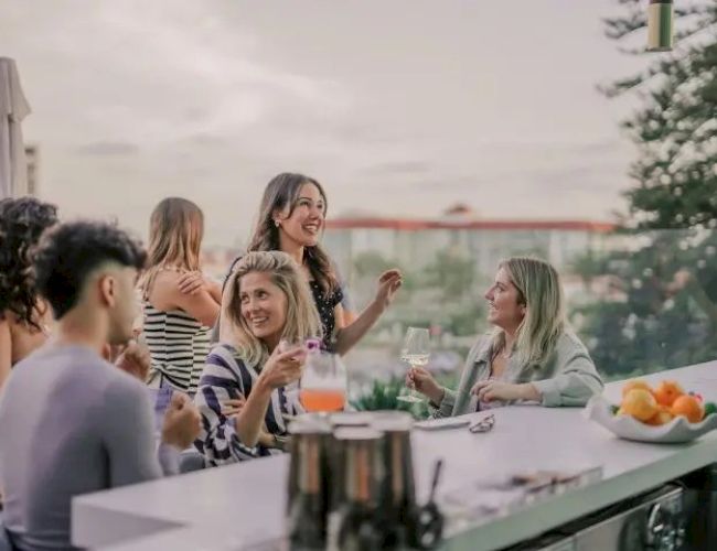 People are enjoying a gathering at an outdoor bar with drinks, fruit, and conversation, set against a scenic background.