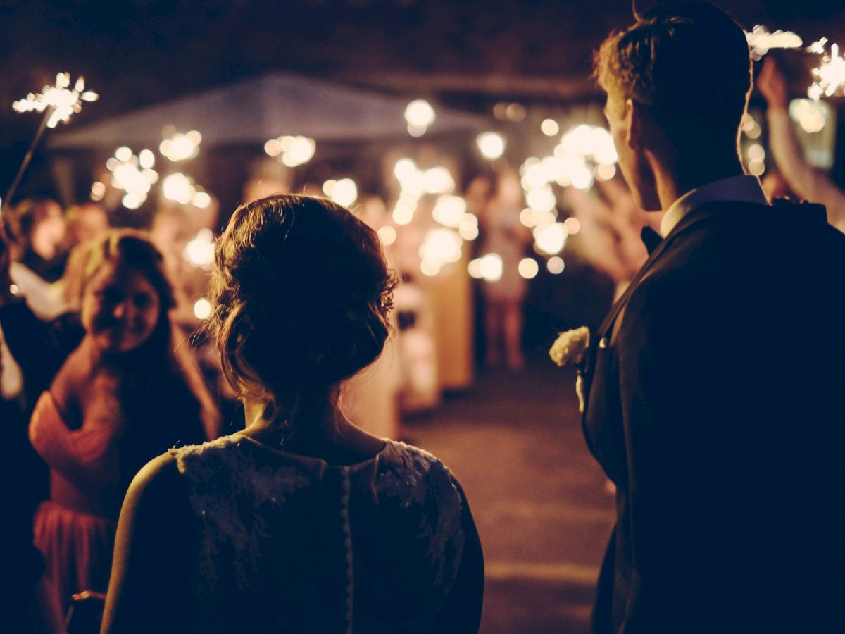 A couple stands with their backs to the camera, surrounded by people holding sparklers in a festive, possibly wedding setting.