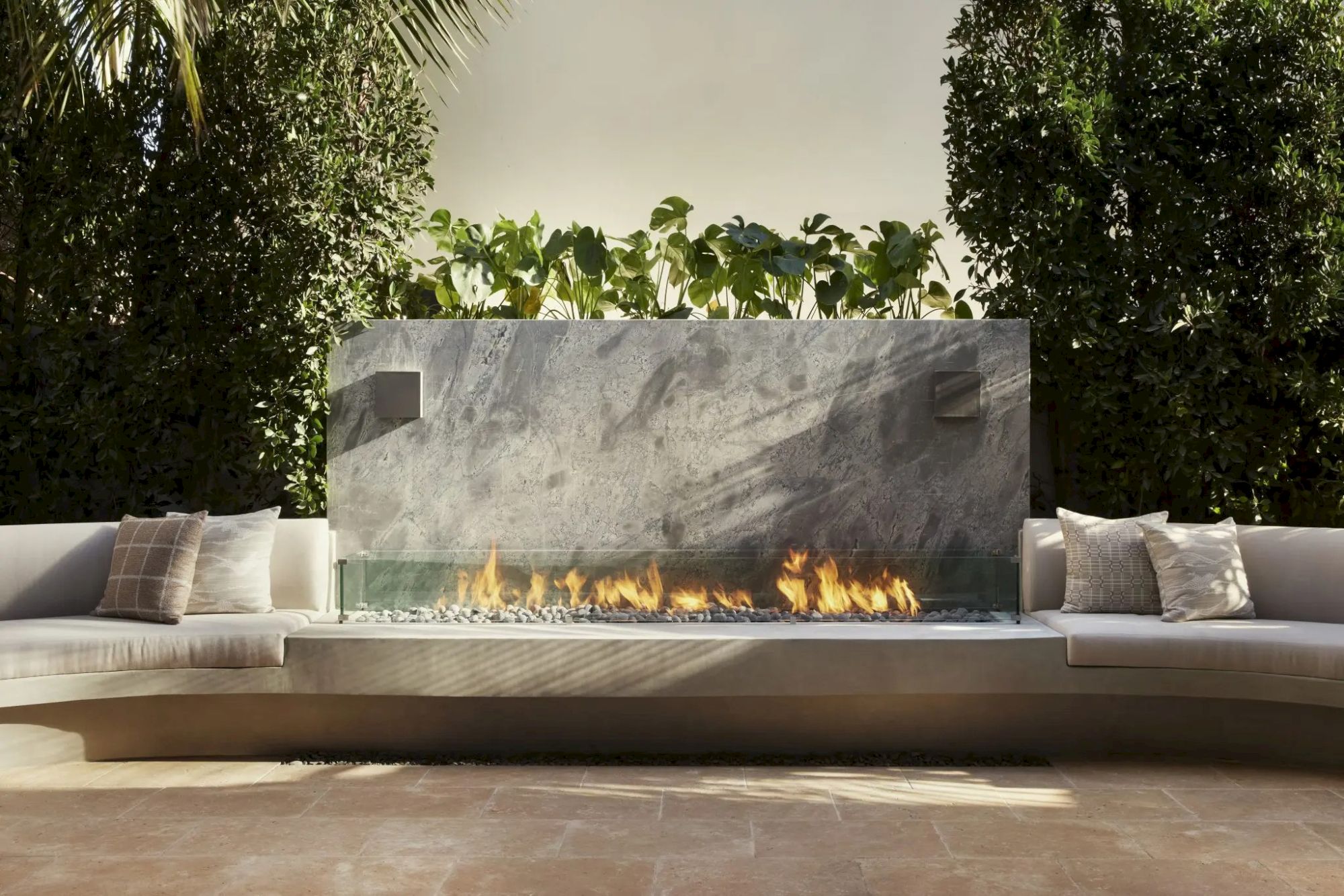 The image shows a modern outdoor seating area with a sleek stone fireplace and greenery backdrop, creating a cozy ambiance.