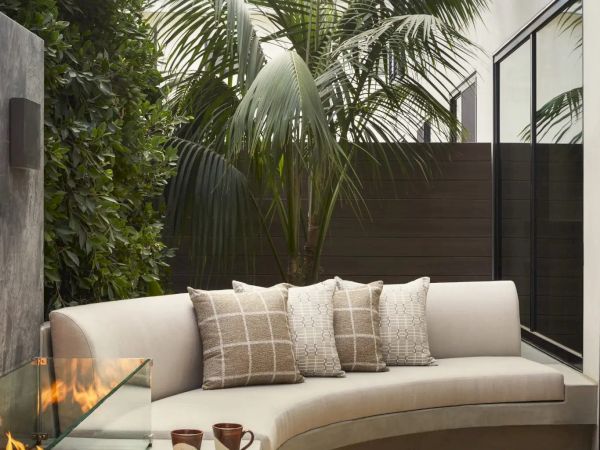A cozy outdoor nook with a curved sofa, palm tree, fire feature, and snacks on a table, next to a modern building.