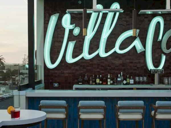 Rooftop bar with neon "Villa" sign, stools at the counter, blue accents, and a small table with a drink, under an open sky.