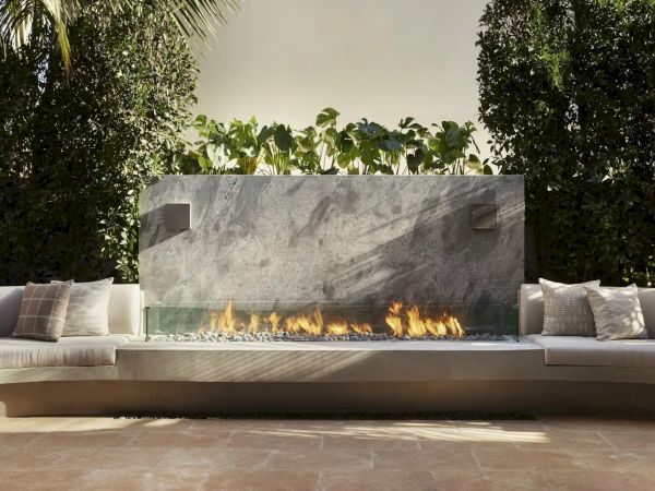 The image shows an outdoor seating area with a curved bench, cushions, a modern fireplace, and lush greenery.