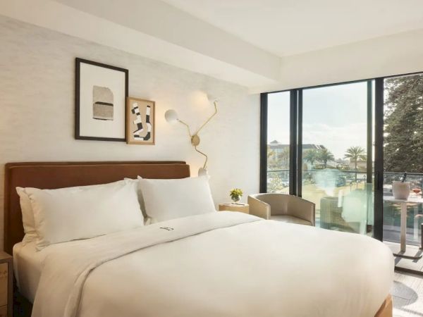 A cozy hotel room with a large bed, modern wall art, soft lighting, and a balcony offering a scenic view of trees and water.