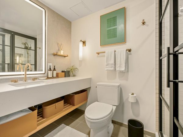 A modern bathroom with a white vanity and sink, large mirror, storage open shelves, wall art, towel rail, toilet, and a glass shower door, all in light, neutral tones.