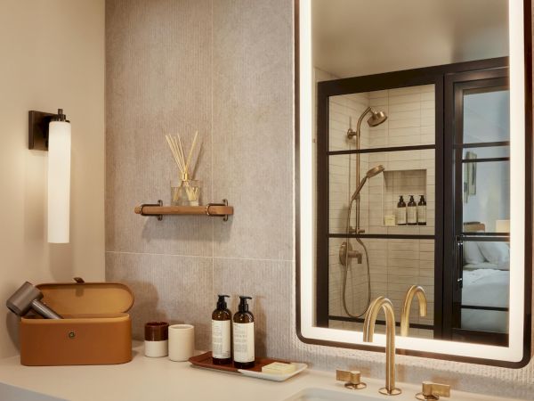 A modern bathroom vanity with a large backlit mirror, gold faucet, double sink, wall shelf, toiletries, and a clean, minimalist layout.