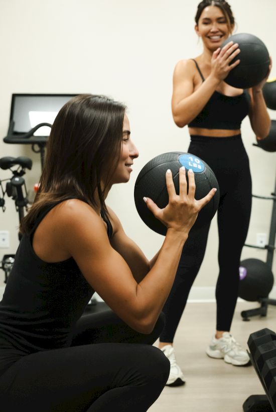 Two women in a gym pass balls between sets: one crouches with a medicine ball, the other smiles standing with another ball beside a workout rack.