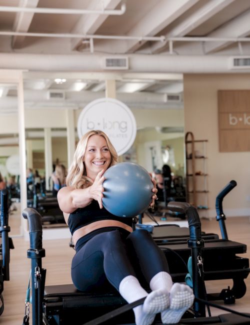 A woman in a gym sits on a Pilates reformer, holding a blue exercise ball and smiling at the camera, mid-workout.