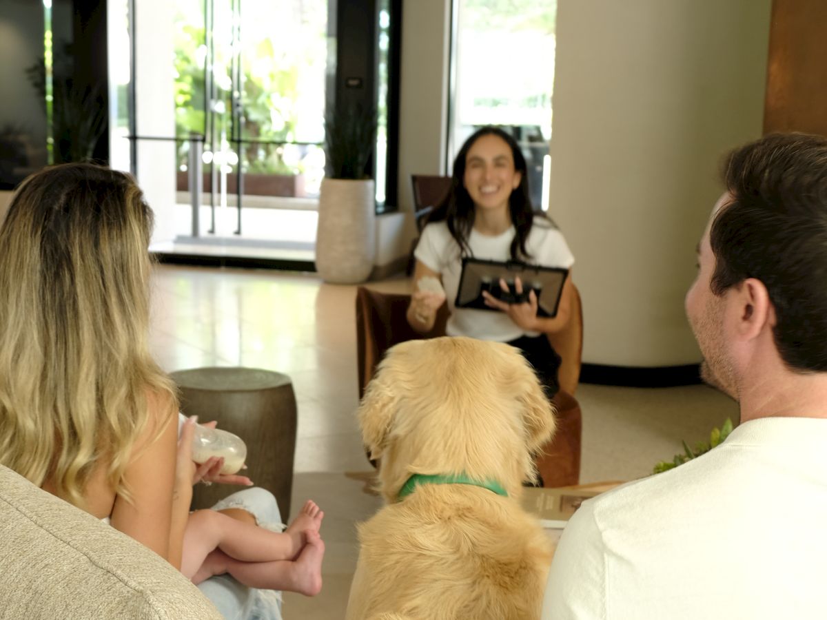 A woman sits on a chair at the front reading from a book to a small group, while a family with a baby and a golden retriever faces her in a cozy lounge. The dog sits between the parents, listening attentively.