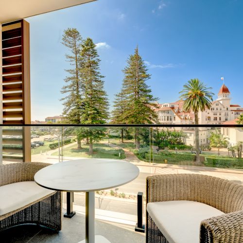 A sunny balcony with wicker chairs and a round table overlooks a green park and distant buildings, including a red-domed structure.