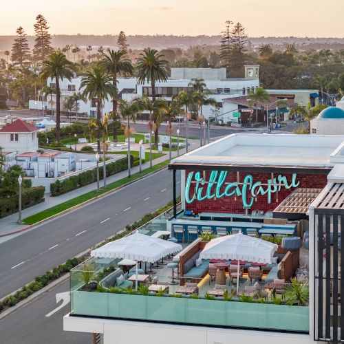 Outdoor rooftop dining at a stylish venue beside a road, palm trees lining the street, with a colorful sign reading "Till Capital" and modern white buildings nearby.