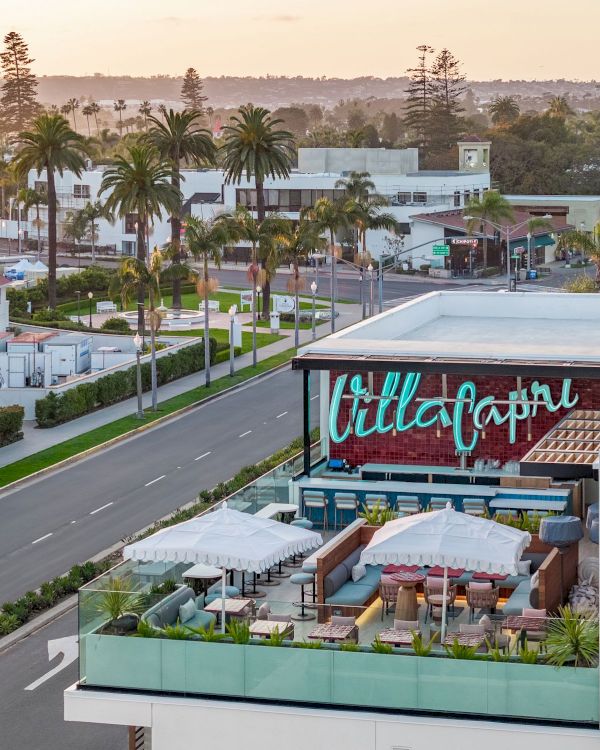 Outdoor rooftop dining at a stylish venue beside a road, palm trees lining the street, with a colorful sign reading "Till Capital" and modern white buildings nearby.