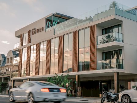 A modern, multi-story hotel with glass balconies and wooden slat accents; a busy street with cars and a motorcycle in front. Top it at 140 characters, always ending the sentence.