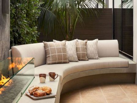 Outdoor seating area with a curved beige sofa, throw pillows, a fire pit, and lush plants in a narrow patio. End.