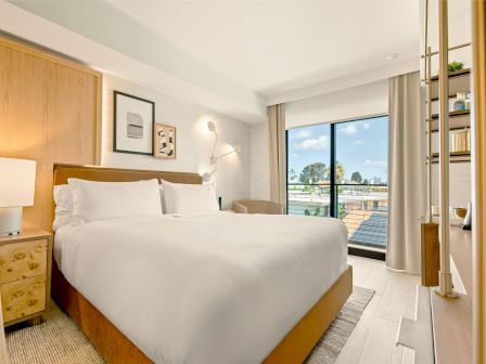 A bright hotel room with a large king bed, nightstands, soft lighting, art on the wall, a seating area, and a balcony view with floor-to-ceiling windows.