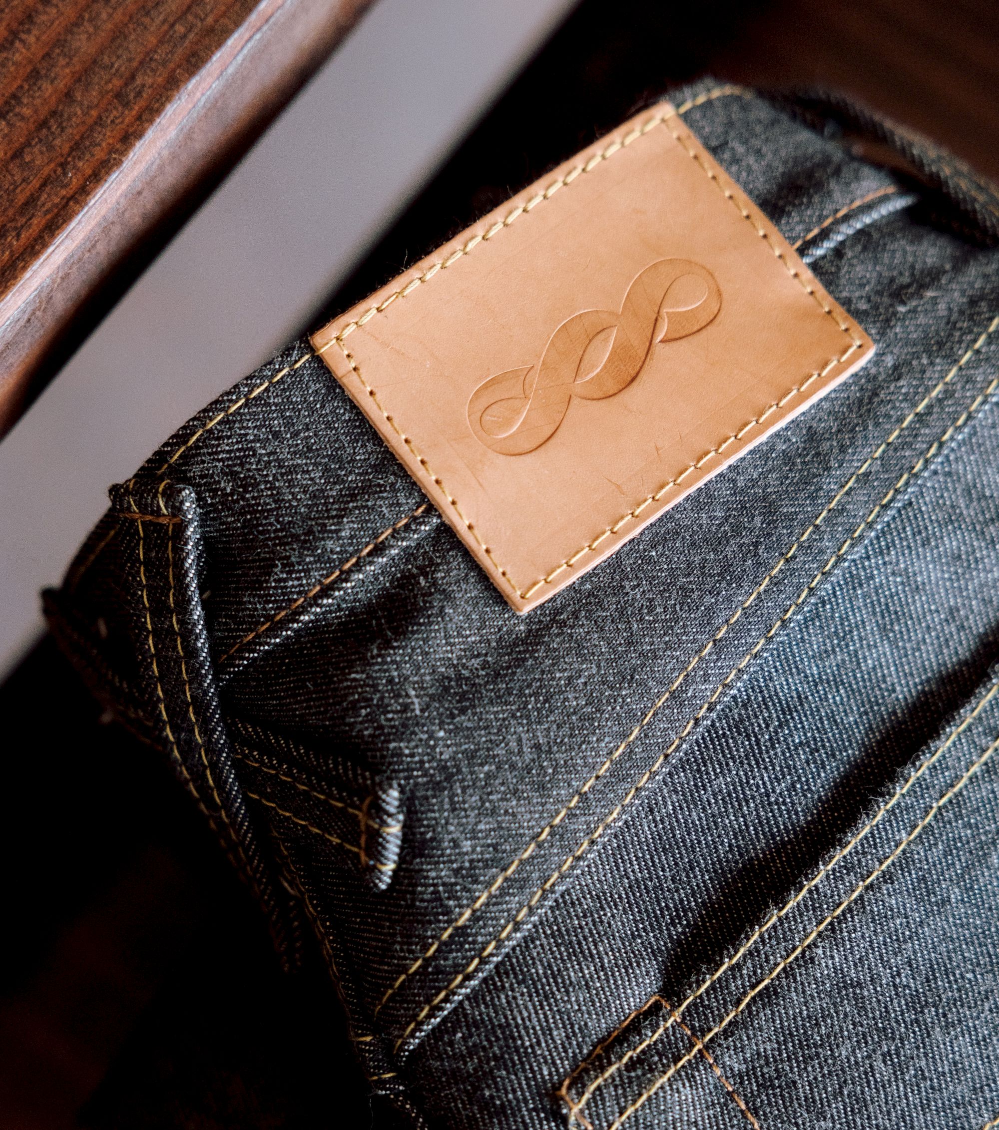 A pair of jeans with a leather patch and stitched pocket, showing a curved seam and a small frayed hole, branded with an embossed infinity symbol.