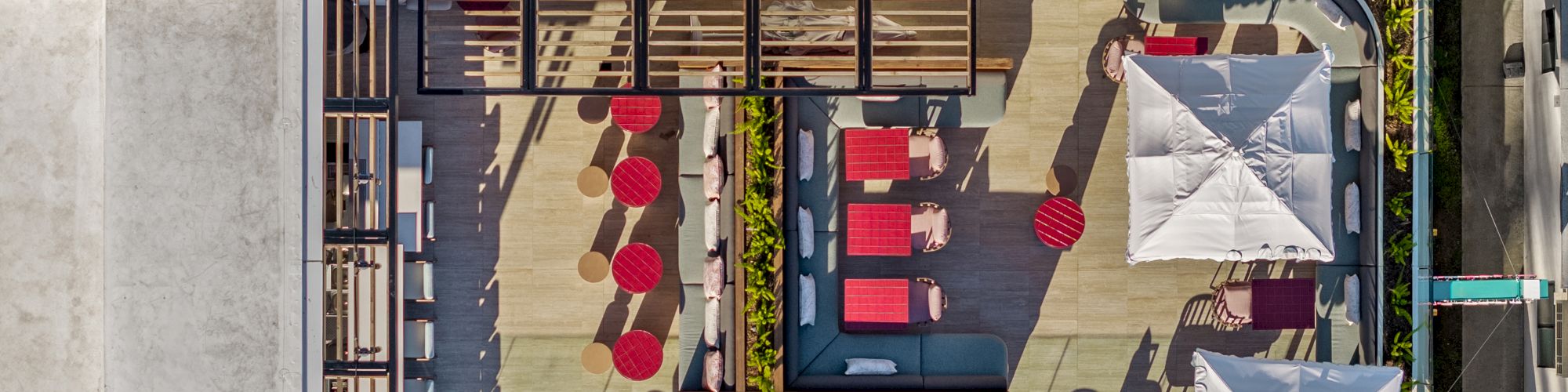 A rooftop terrace with red seating, a central lounge area, sun umbrellas, bar seating along the edge, and potted plants surrounding a stylish outdoor space.