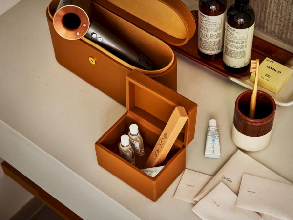 A neatly arranged vanity with a tan leather case, small box of toiletries, tubes, bottles, a toothbrush cup, and skincare sachets on a white counter.