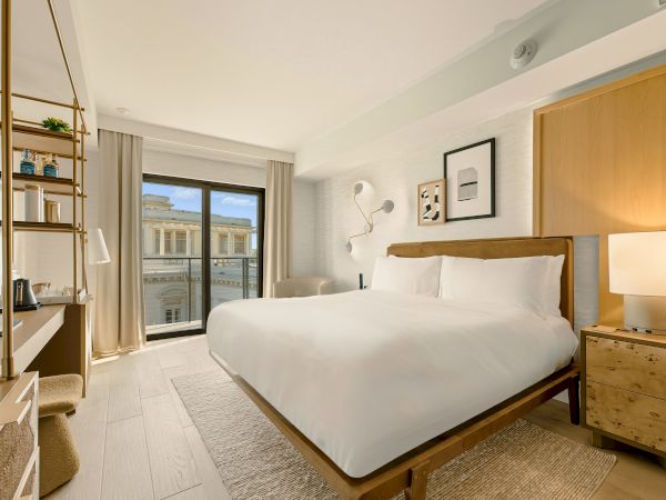 A bright hotel room with a large white bed, wooden headboard, nightstands, lamps, a desk area, and a sliding glass door to a balcony. End.