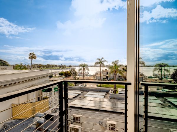 A sunny coastal view from a balcony showing modern buildings, palm trees, clear blue sky, and a glimpse of the ocean in the distance.