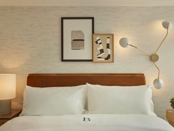 A modern hotel bedroom with a white bed, brown headboard, soft lamps, abstract wall art, and a stylish wall-mounted light fixture, exactly as shown.