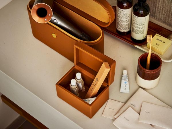 A stylish travel kit with a tan leather case, a small open box containing bottles and tools, tubes, and several neatly arranged sachets on a counter.